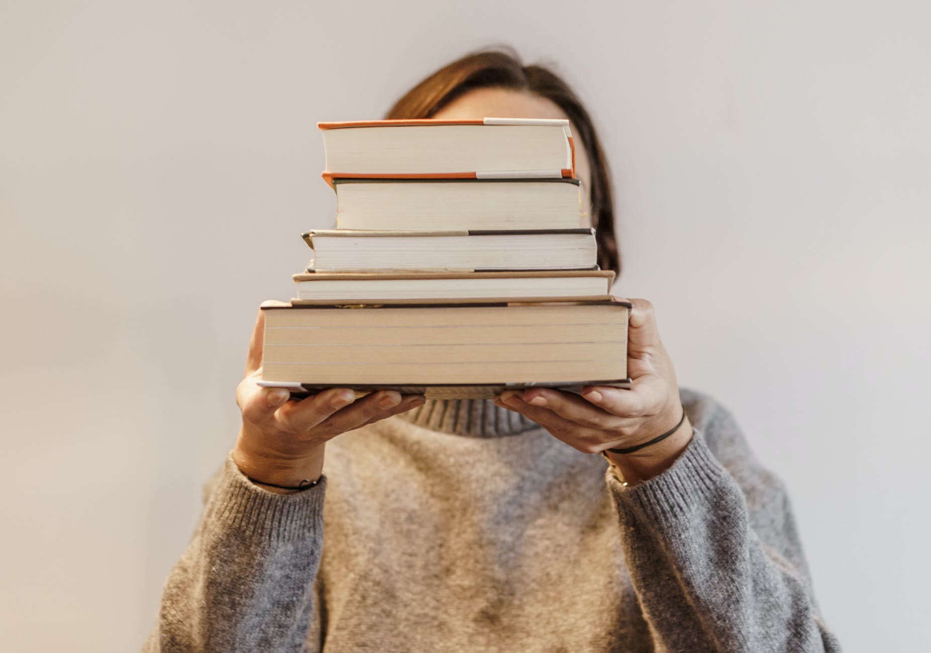 person in gray sweater carrying a stack of hardbound books